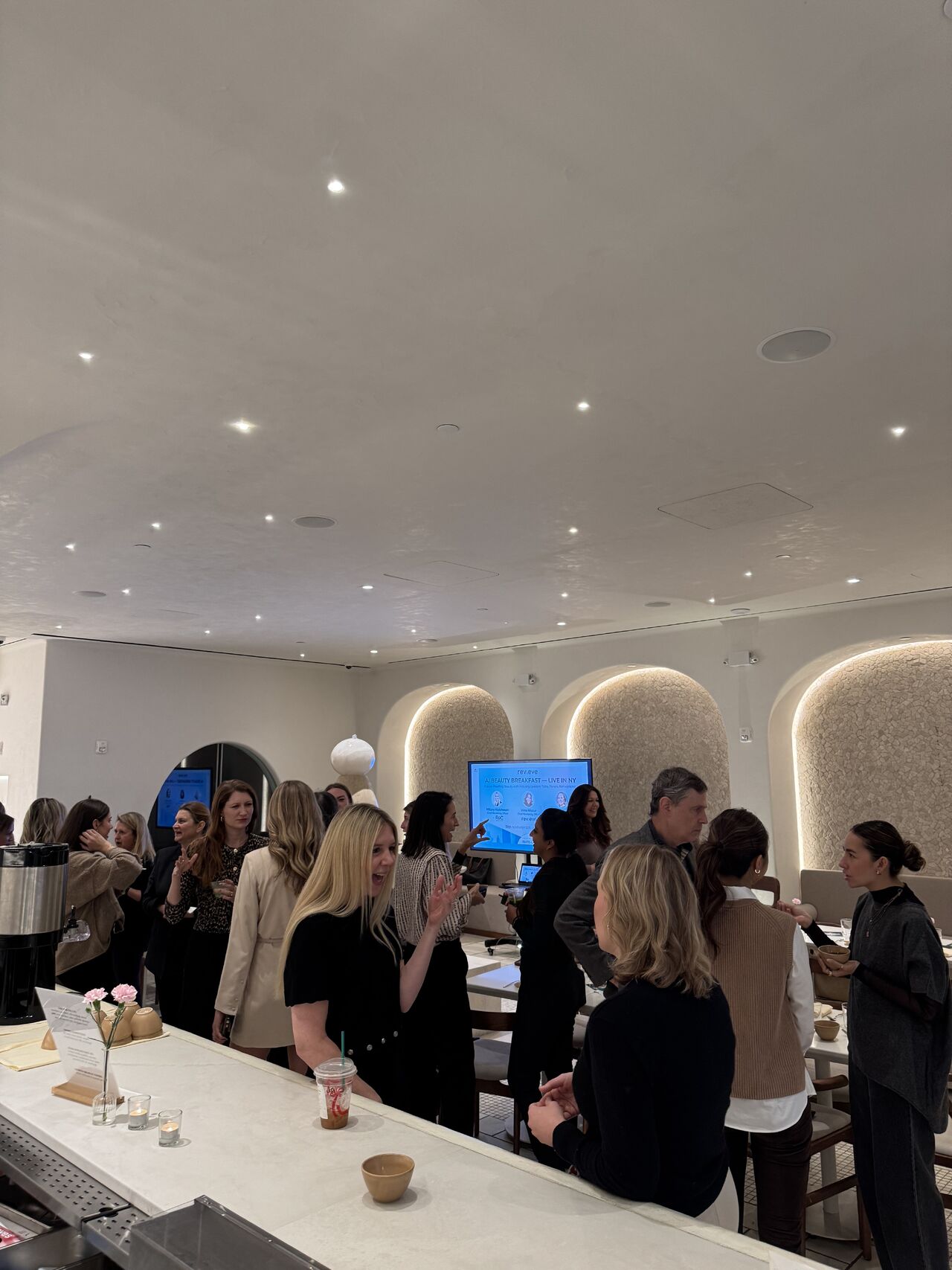 Guests networking at Revieve's AI Beauty Breakfast in New York, with a welcome screen reading “AI Beauty Breakfast — Live in NY”